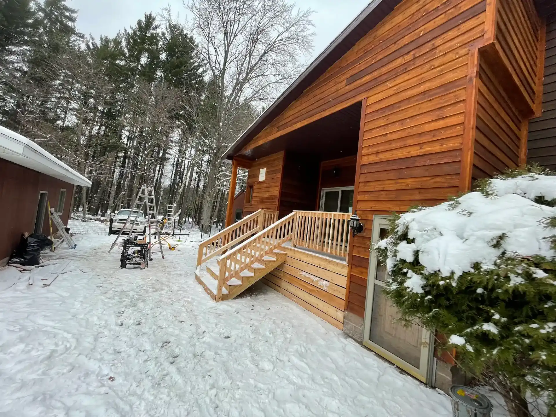 Completed exterior renovation with cedar siding, new porch stairs, and railings on a wooded property in winter in Marquette, Michigan