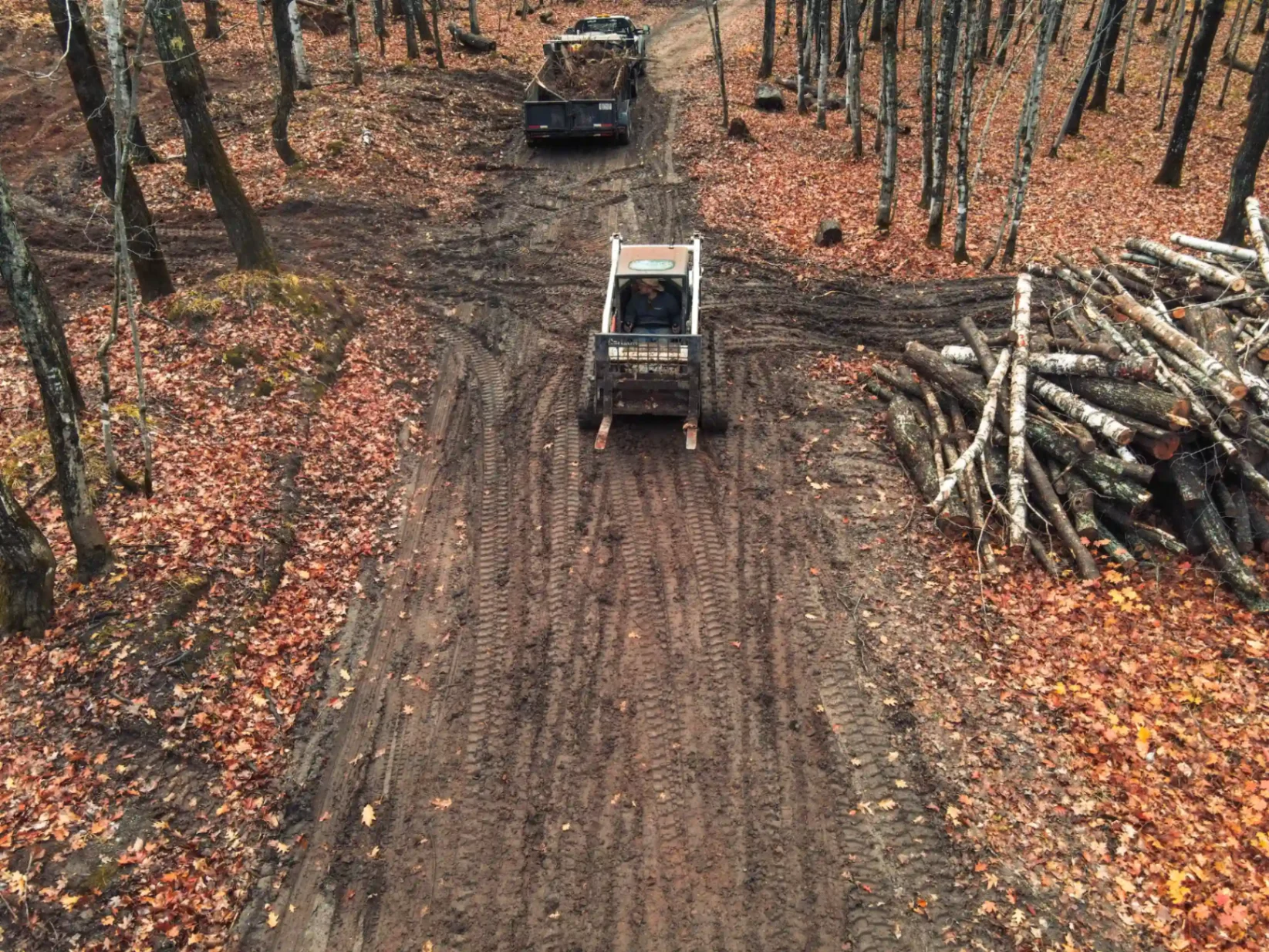Aerial view of skid steer grading a private road through hardwood forest with stacked birch logs in Marquette County, Michigan