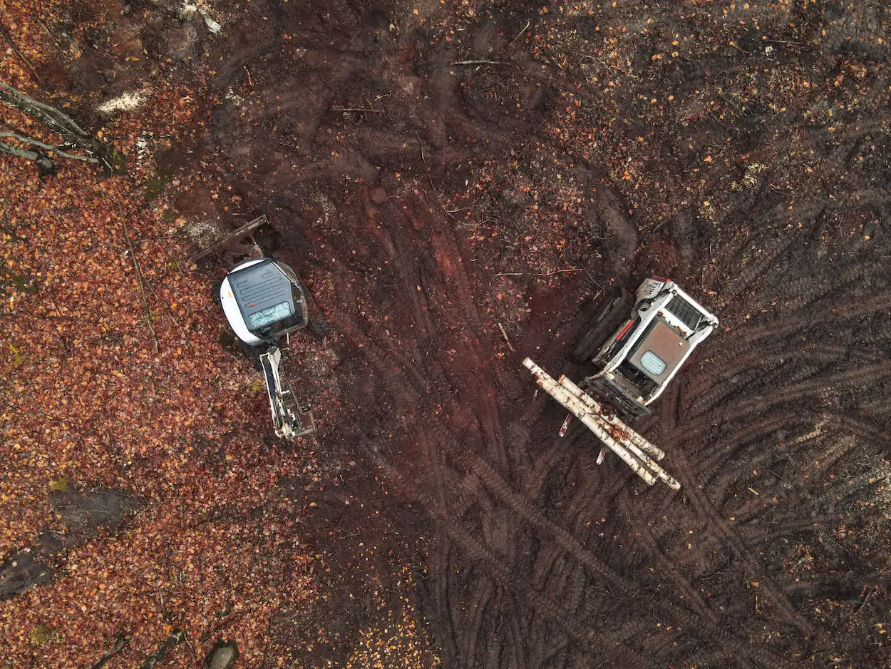 Aerial view of excavator and skid steer clearing a wooded lot with red clay soil and birch logs in Marquette County, Michigan